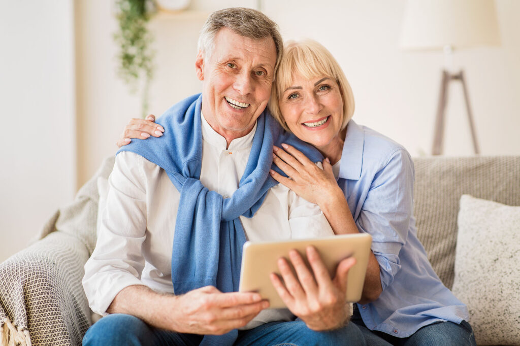 Excited mature couple with digital tablet smiling to camera, woman embracing husband